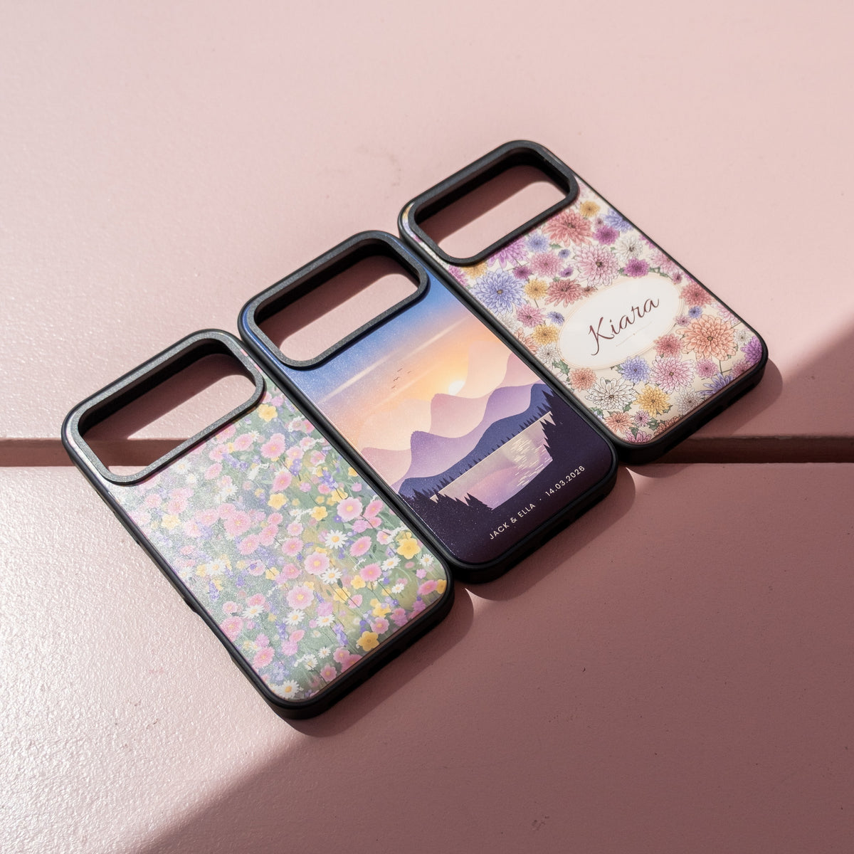 Three custom skin MagSafe iPhone cases on a pink wall: wildflower meadow, mountain sunset with names, and a floral design with the name Kiara