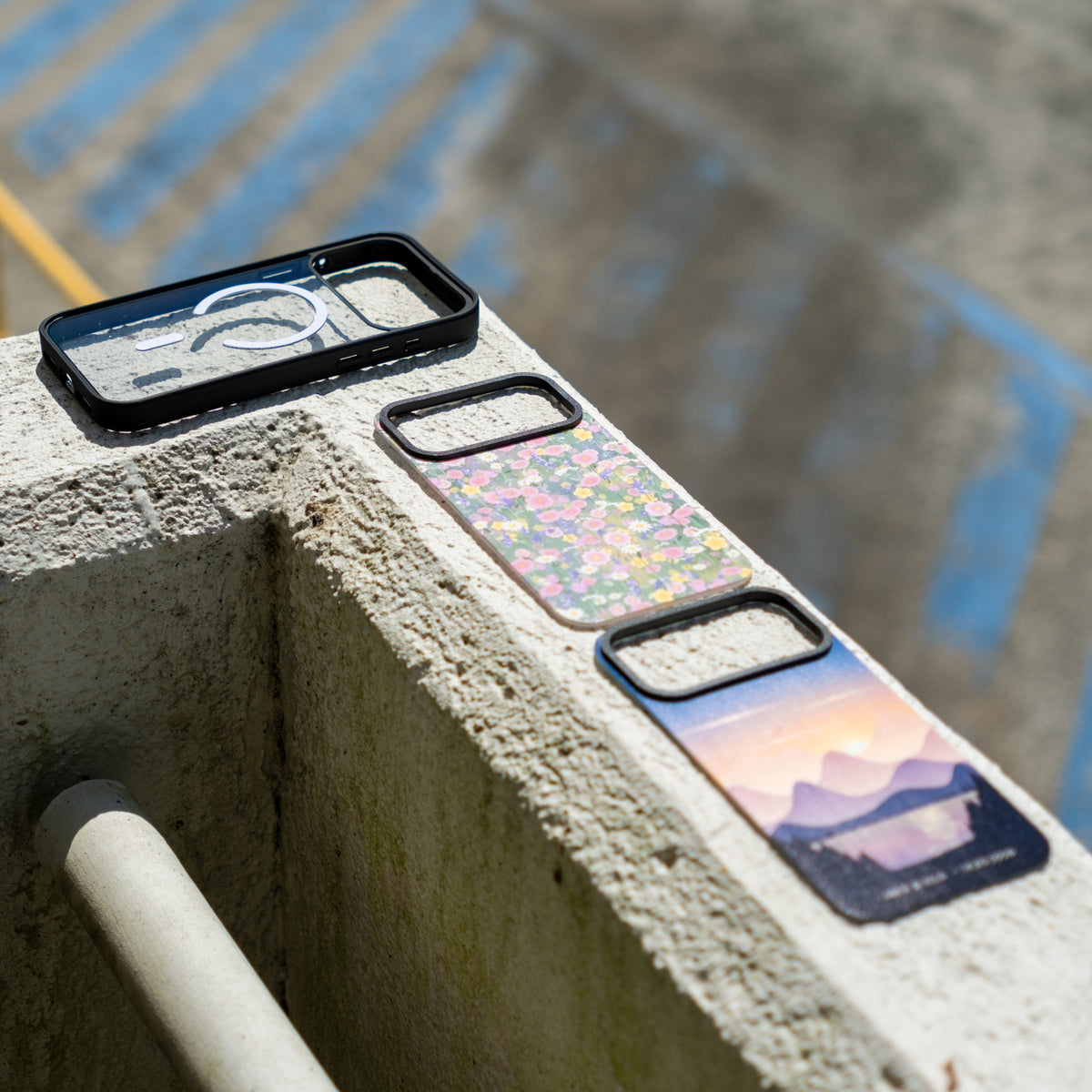 Custom skin MagSafe iPhone cases on a concrete ledge outdoors: clear case, wildflower skin and mountain sunset skin