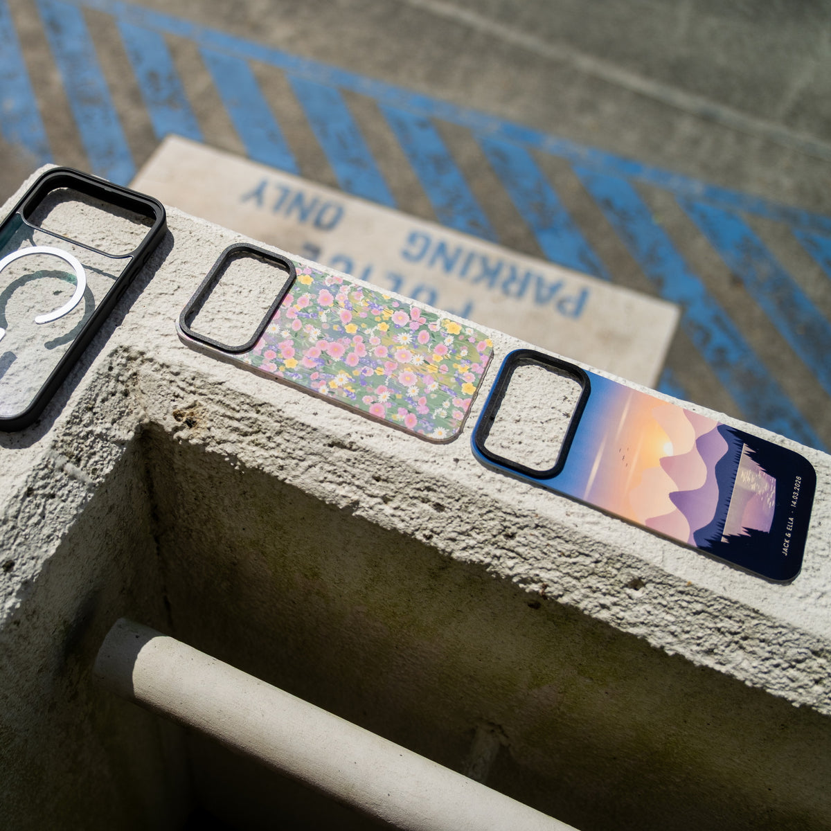 Three custom skin MagSafe iPhone cases on a concrete ledge outdoors, wildflower and mountain sunset designs