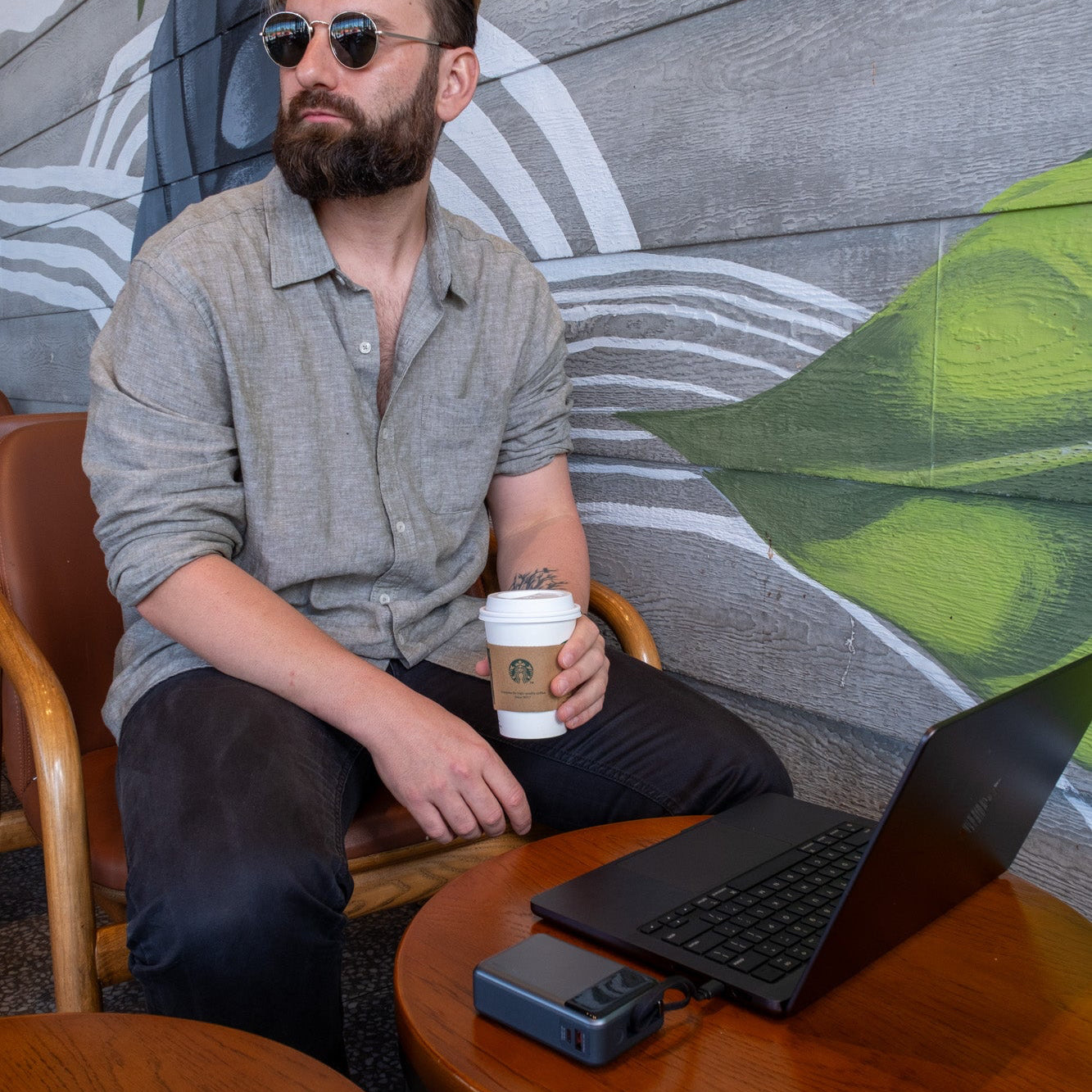 Man sitting at a table with a laptop, power bank and coffee cup, against a wall with green leaf designs.