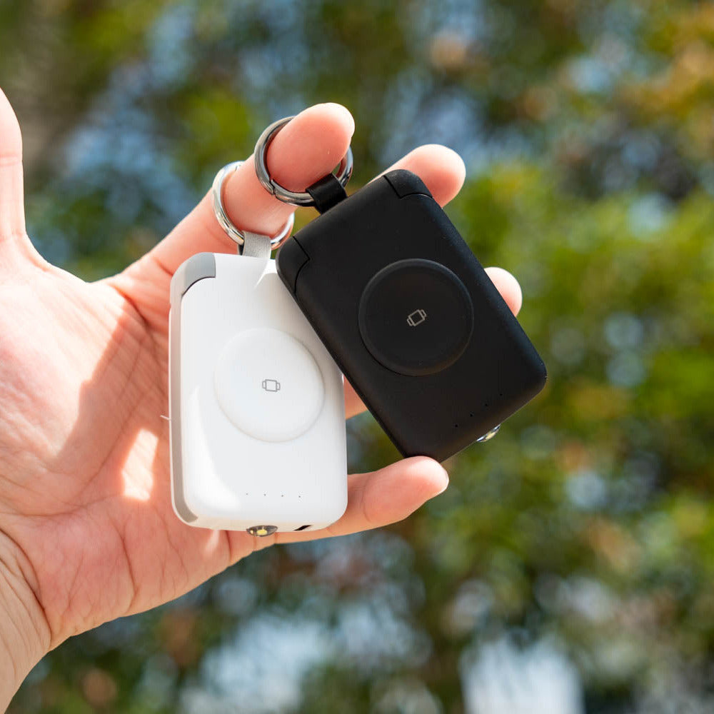 Hand holding black and white PTC WatchGo 2000 power banks by their keyrings outdoors