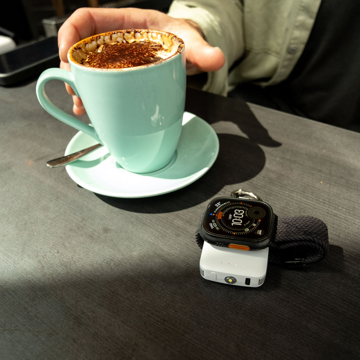 Apple Watch Ultra charging on a white PTC WatchGo 2000 on a cafe table beside a coffee