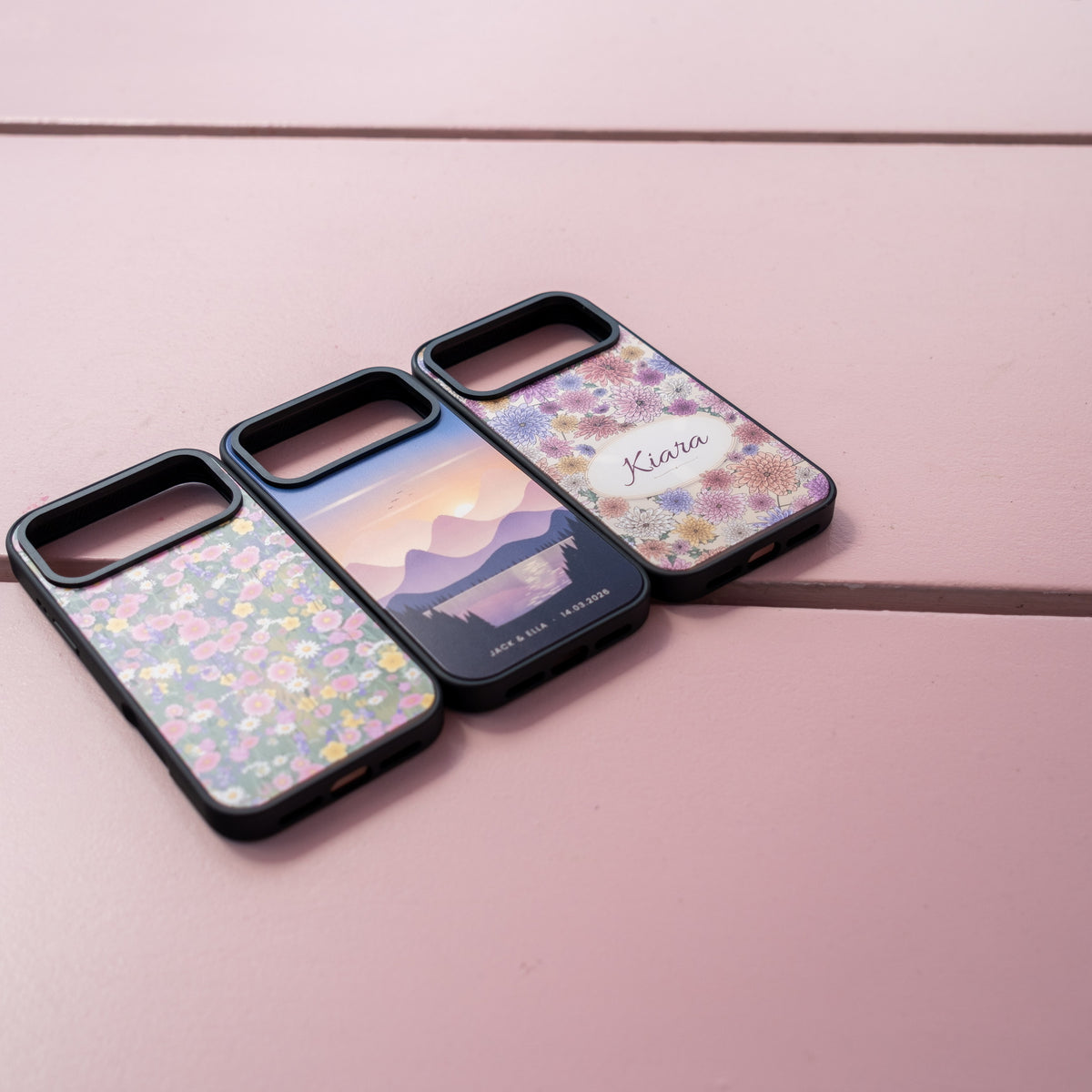 Custom skin MagSafe iPhone cases lined up on a pink ledge: wildflower meadow, mountain sunset with names and date, and a Kiara floral name design